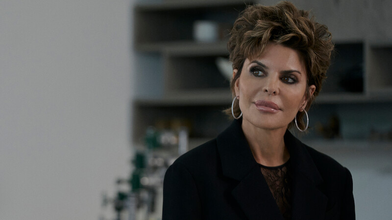 Woman with short styled hair and hoop earrings wearing a black blazer in a modern indoor setting with shelves and bottles blurred in the background, looking thoughtfully to the side.