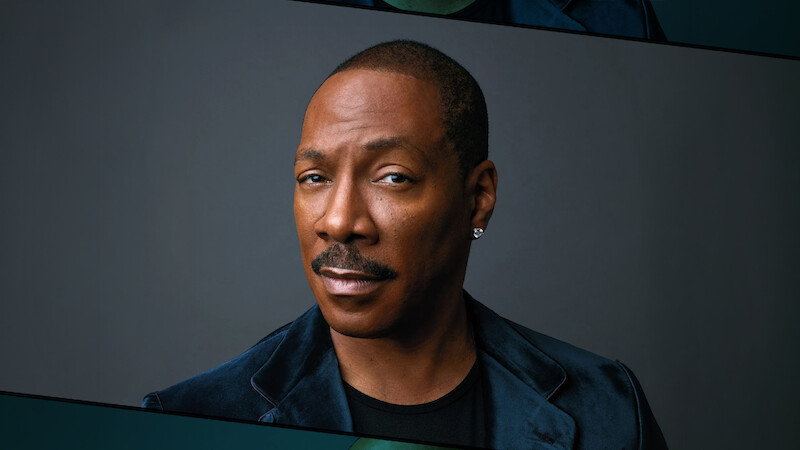 Eddie Murphy with short hair, mustache, wearing a dark velvet jacket and diamond earring, poses against a plain dark background, with a serious and confident expression. Studio lighting highlights his face.