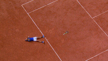 A tennis player lies on a clay court next to a tennis racquet, possibly celebrating or exhausted, viewed from above. The scene captures the lines and earthy texture of the tennis court.