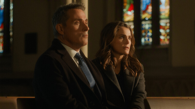 Two people sitting on a bench inside a dimly lit church, stained glass windows in the background, both wearing formal clothing and looking ahead with thoughtful expressions.