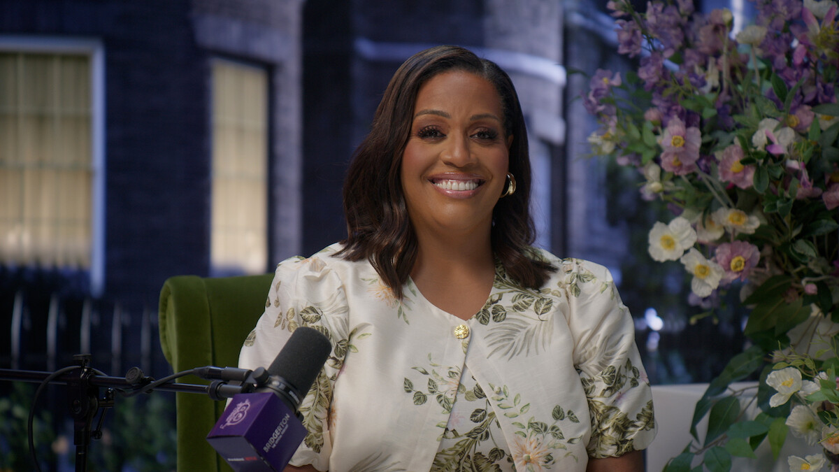 Woman smiling at a podcast setup with a microphone in front, sitting indoors next to a large floral arrangement, with blurred windows and greenery in the background.