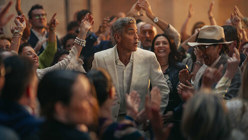 A man in a white suit stands surrounded by a lively crowd at a festive gathering, with smiling faces and raised hands creating a joyful, celebratory atmosphere.