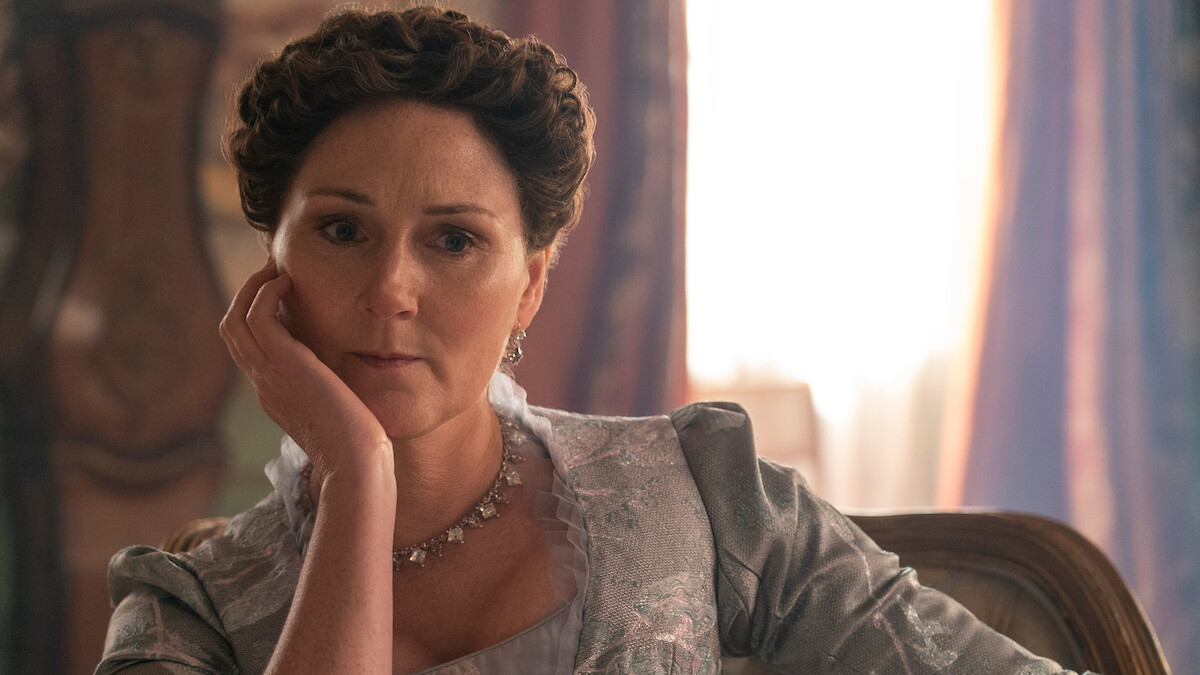 A woman with styled brown hair and vintage clothing sits indoors on a chair, resting her head on her hand, with soft natural light and ornate décor in the background, evoking a historical or period setting.
