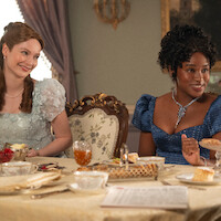 Two women in elegant period dresses sit at a lavishly set dining table in a richly decorated room, enjoying tea and desserts, with ornate chairs and a soft natural light coming through curtained windows.