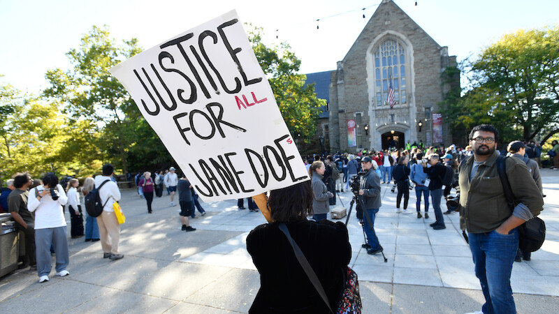Person holds "Justice for Jane Doe" sign at outdoor protest in front of a large stone building, with a crowd of people gathered around under a sunny sky with trees in the background.