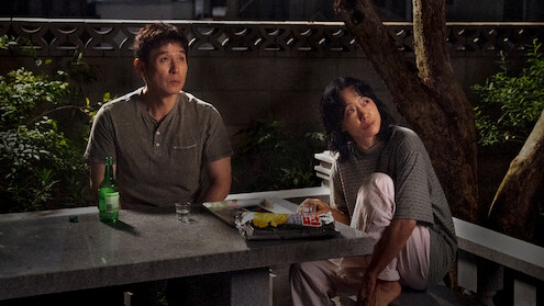 Two people sit at a stone table outdoors at night, surrounded by trees and plants, with snacks, a bottle, and a glass on the table, looking upward as if observing something in the sky.