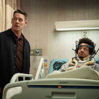 A man wearing a dark jacket stands beside a hospital bed where another man lies with a head brace and neck support, surrounded by medical equipment in a hospital room, patient appears injured.