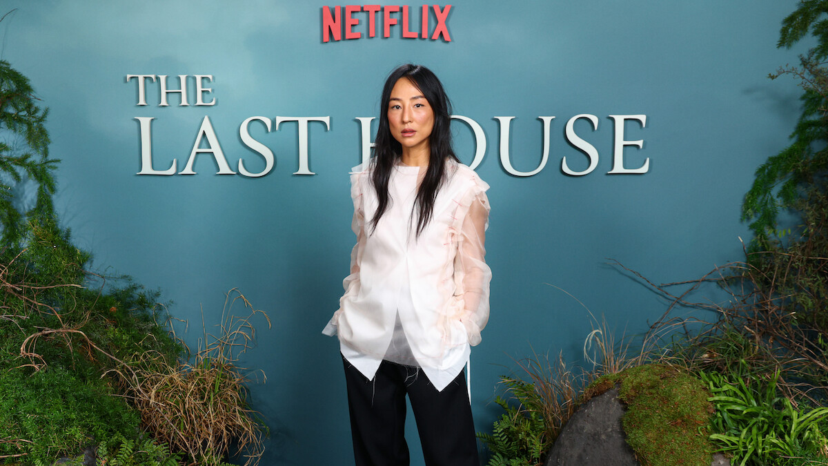 Greta Lee stands in front of a blue backdrop with "Netflix" and "The Last House" text, surrounded by greenery and natural elements, at a promotional event or premiere.