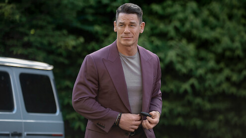Man in a mauve suit jacket and gray shirt stands outdoors near a silver van, with a background of dense green foliage, holding an object and looking slightly to the side.