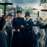 A woman in Victorian-era clothing, handcuffed and escorted by a police officer, stands amid an angry protest crowd holding signs and torches outdoors, suggesting a historical or dramatic arrest scene.
