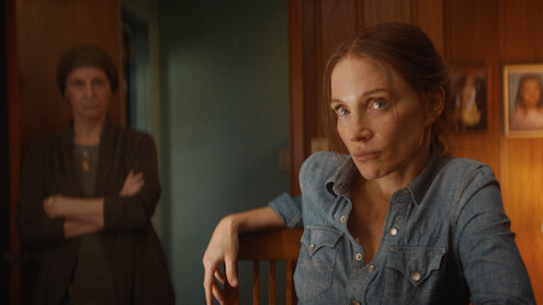 Woman in a denim shirt sits at a wooden table in a warmly lit room, looking serious. Another woman stands in the background with arms crossed. Wooden panel walls and family photos create a cozy, tense atmosphere.