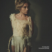 Woman with wavy blonde hair in an artistic, fringed cream top stands against a dark, plain background, looking to the side; moody editorial photo with dramatic lighting. Tudum Magazine logo in the corner.