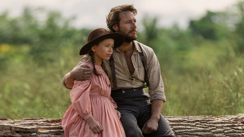 A man and a young girl in old-fashioned clothing sit on a large log outdoors. The man has his arm around the girl, and they both look thoughtfully into the distance, surrounded by greenery and soft natural light.