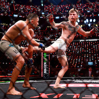 Two mixed martial artists fighting in a cage during a professional match, with one delivering a kick to the other's leg. The arena is filled with lights and the audience is visible in the background.