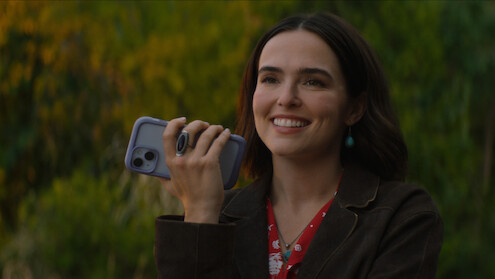 A woman smiling outdoors, holding a smartphone in her hand, wearing a brown jacket and red floral blouse, with green blurred bushes and trees in the background on a sunny day.