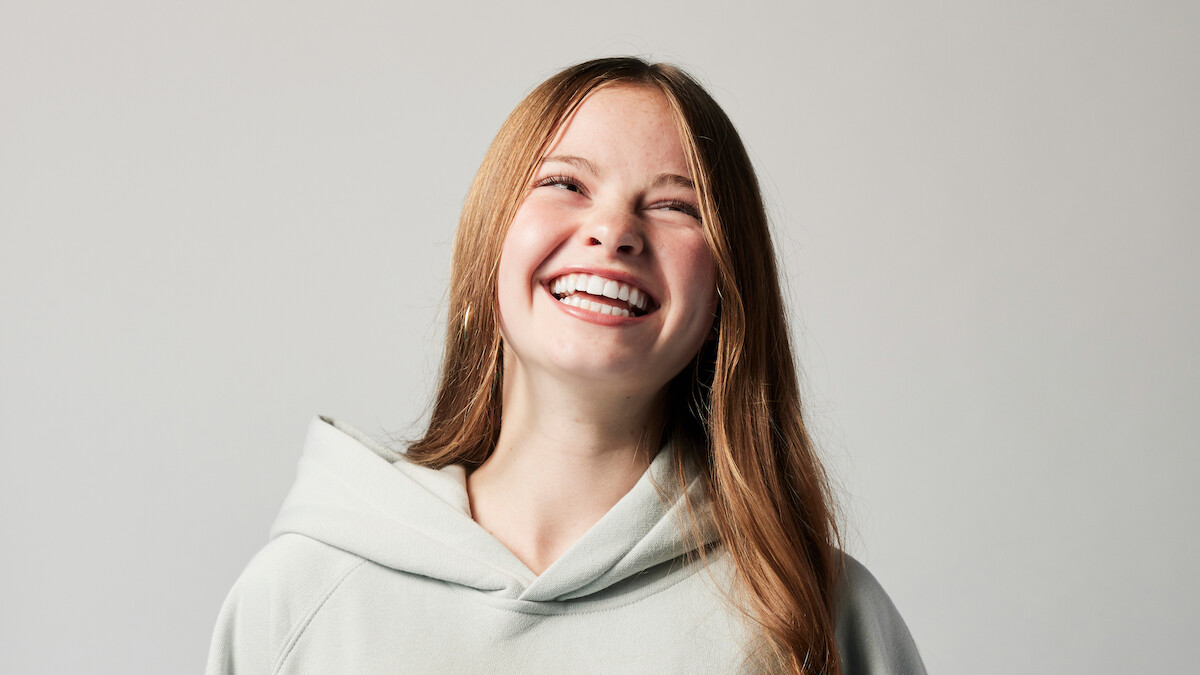 Smiling young woman with long hair wearing a light hoodie, standing against a plain, light-colored background in a studio setting.