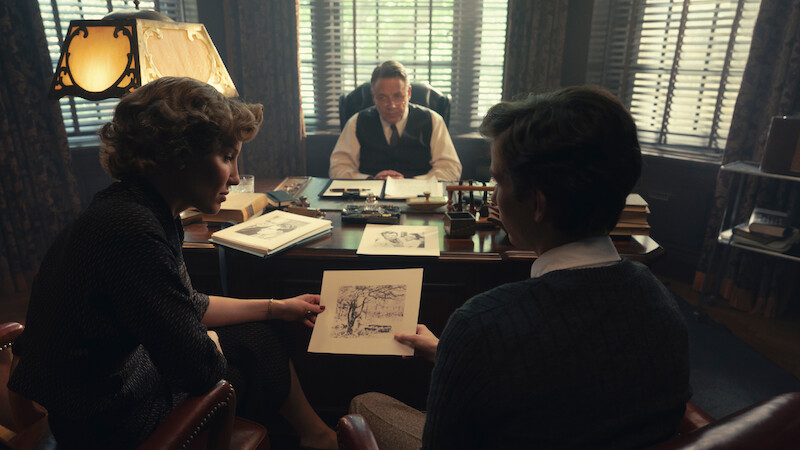 Two people sit across from a man at a desk in a dimly lit office, discussing a drawing. The office has large windows with blinds, books, and papers scattered on the desk, creating a serious, professional atmosphere.