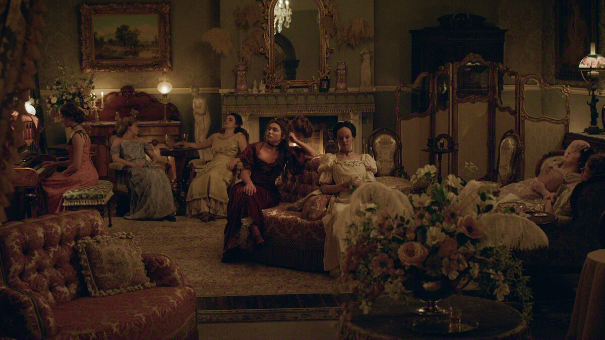 Women in elegant period dresses sit and lounge in a richly decorated parlor with vintage furniture, ornate decor, flowers, and a lit fireplace, creating a warm, historical atmosphere.