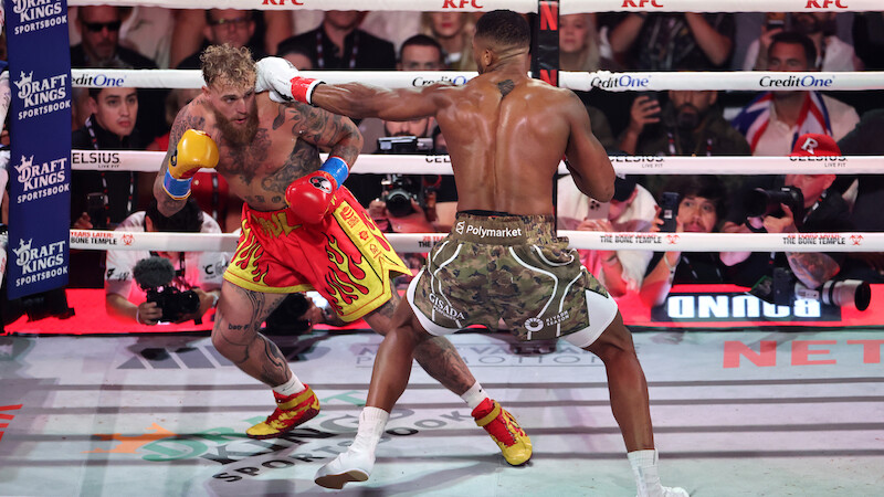 Jake Paul and Anthony Joshua fight in a brightly lit boxing ring, surrounded by ropes and an audience, with camera crews and sponsors visible in the background.