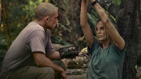 A man kneels beside a woman tied to a tree in a dense forest, holding a canteen. Both look dirty and tense, suggesting a survival or hostage situation in a rugged, outdoor setting.
