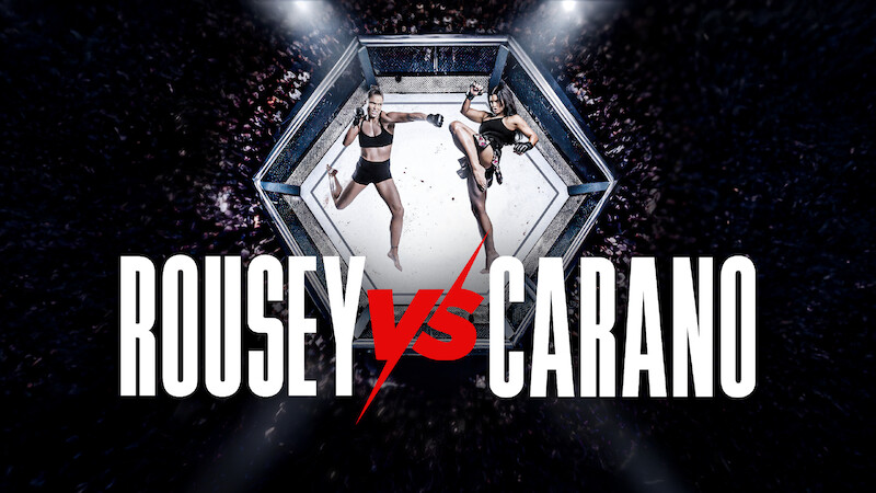 Two female fighters facing off in a mixed martial arts octagon cage, surrounded by a cheering crowd, with large text reading Rousey vs Carano in front.