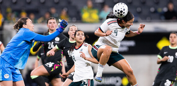 Women's soccer match in a stadium, goalkeeper in blue reaches for the ball as player in white and green jumps to head it, teammates and opponents in action, focused expressions, blurred spectators in background.
