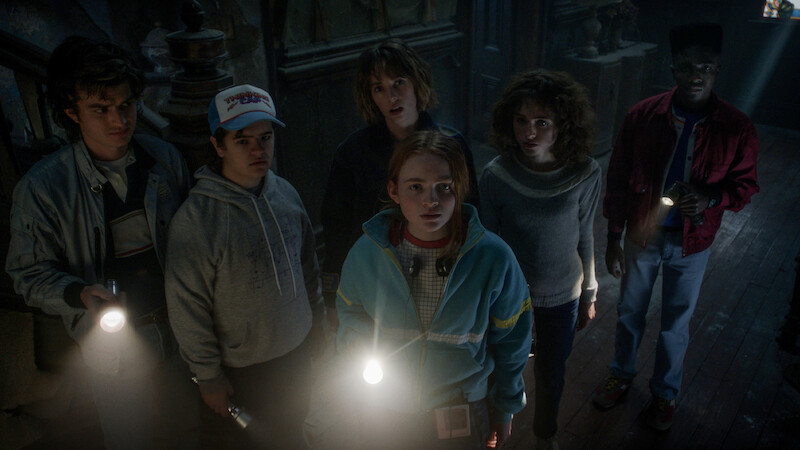 Six teenagers stand close together in a dark, old building holding flashlights, looking up with serious and tense expressions, creating a suspenseful and mysterious mood.
