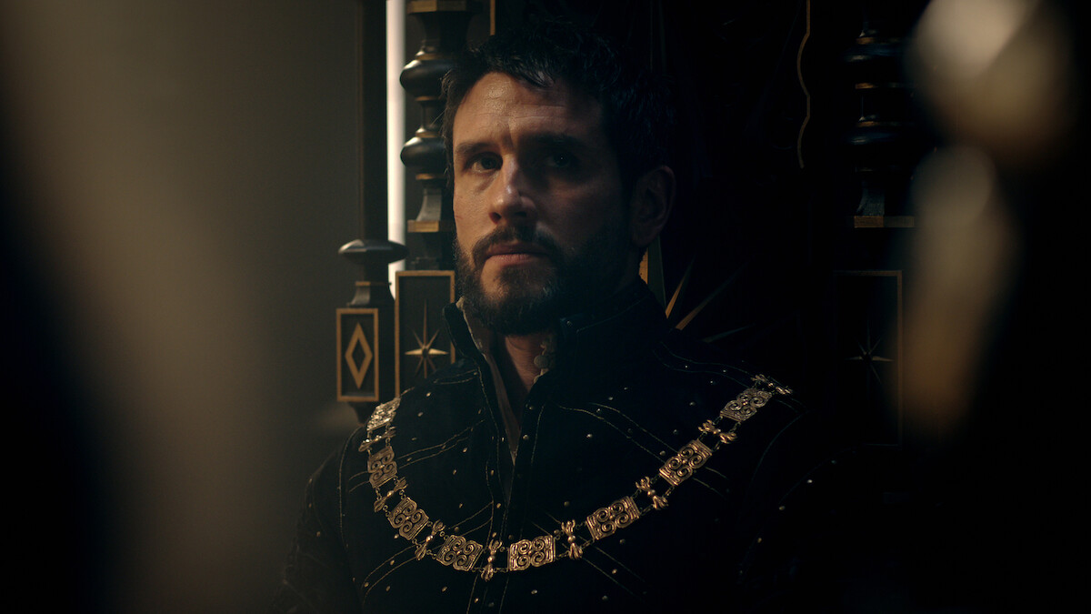 A solemn man in elaborate medieval attire with a gold chain sits on a dark, ornate throne in a dimly lit, regal setting, creating a moody, contemplative atmosphere.