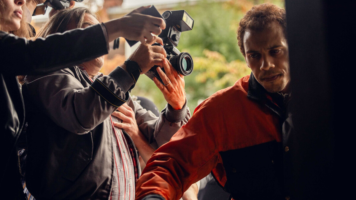 A man in a red and black jacket tries to move through a crowd outside as people with cameras and microphones surround him, suggesting a media or paparazzi situation.