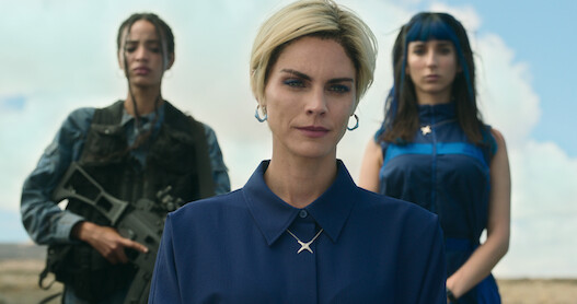 A woman stands facing the camera while to others stand behind her — one of whom is holding a rifle.