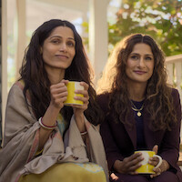 Two women sitting on a porch, holding yellow mugs, dressed in cozy and semi-formal clothes, smiling and enjoying a peaceful outdoor moment with sunlight and greenery in the background.