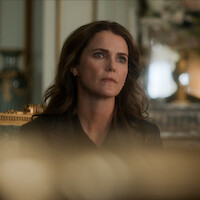 A woman with wavy brown hair sits indoors in a formal, ornate room with gold accents, appearing thoughtful or concerned. The background is softly blurred, highlighting her facial expression and the elegant setting.