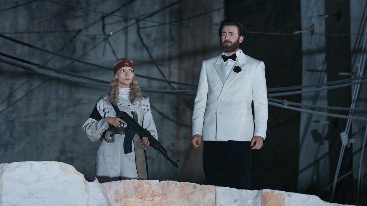 A woman holding a rifle stands next to a bearded man in a white tuxedo jacket. They are in an industrial setting with exposed wires and a rough concrete wall behind them.