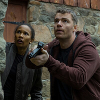 Two people outside by a stone wall, looking alert and cautious. One holds a handgun, both in casual clothing, suggesting a tense or dangerous situation, possibly investigating or preparing for action.