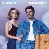 Two people stand closely together, one in a dark tank top and the other in a denim jacket, against a pastel sky backdrop with the words "TUDUM MAGAZINE" above them. An orange patterned chair is partially visible in the foreground.