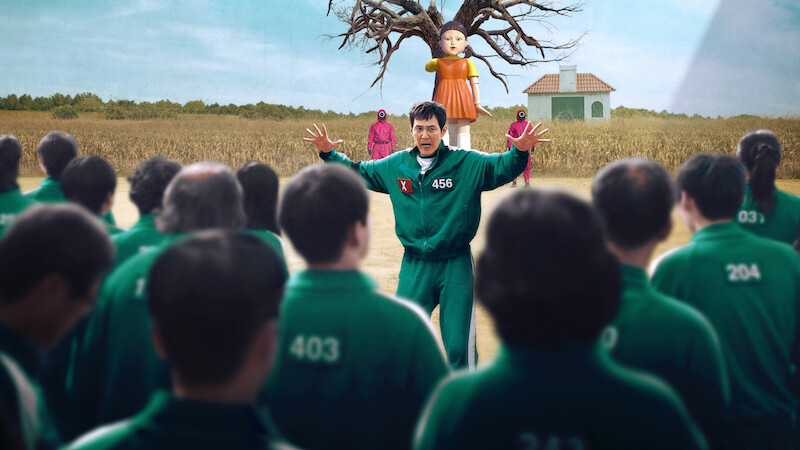 Group in green tracksuits faces man #456 with arms raised in a field. A giant doll, tree, and pink-clad guards are in the background near a small building.