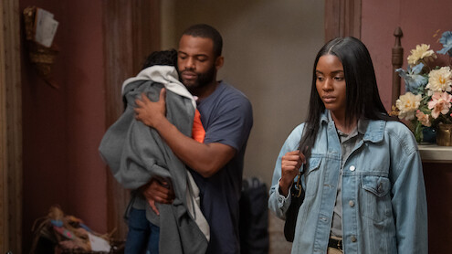 A man holding a child stands beside a woman in a denim jacket. They are indoors in a warmly lit room with flowers on a shelf and decorative objects in the background. The mood appears serious or contemplative.