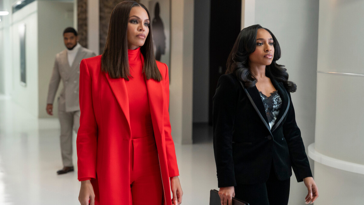 Crystle Stewart as Mallory and Taylor Polidore Williams as Kimmie in business attire, one in a red suit and the other in a black suit, walk confidently down a modern office hallway, with a man in a grey suit walking behind them.