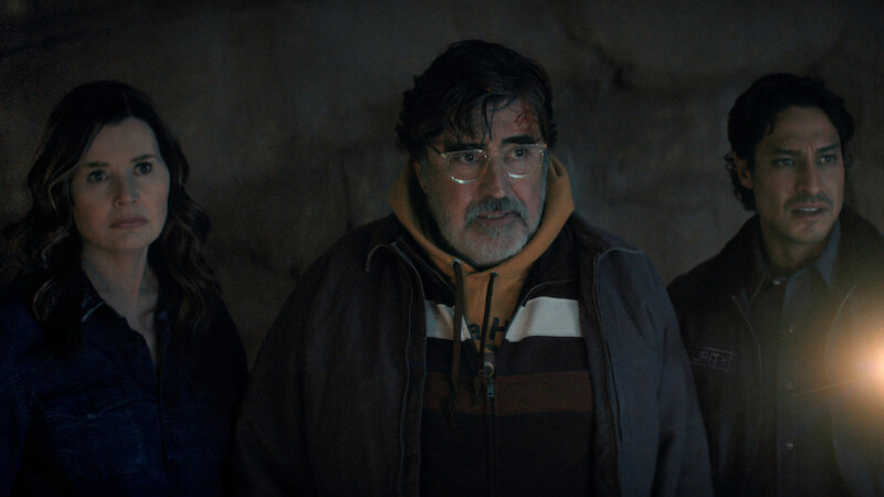 Three people stand together in a dimly lit, cave-like environment, looking ahead with serious expressions. One person holds a light source, adding a dramatic glow to the mysterious scene.
