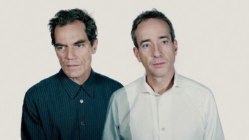 Two men stand close together against a plain light background, one in a dark striped shirt and the other in a white shirt, both wearing serious expressions, creating a somber and introspective mood.