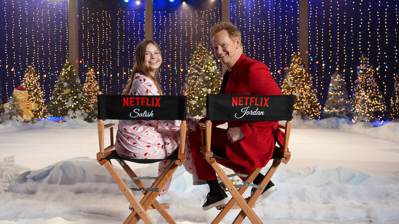 Two people smiling while sitting on director’s chairs with Netflix logos in a festive winter set decorated with snow, Christmas trees, twinkling lights, and holiday decorations, creating a cheerful holiday atmosphere.