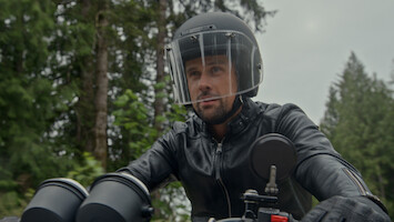 Um homem de capacete preto e jaqueta de couro pilotando uma moto em uma área florestal, com árvores e céu nublado ao fundo.