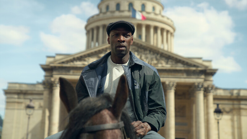 A man in a dark jacket and cap sits on a horse in front of a grand historic building with columns and a French flag, under a partly cloudy sky.