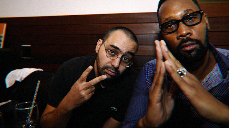 Two men sitting indoors at a table, one making a peace sign and the other with hands pressed together, both wearing glasses in a dimly-lit setting with wooden paneling in the background.