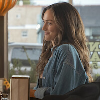 Minka Kelly as Quinn with long brown hair in a denim shirt sits at a table in a cozy diner, smiling, with large windows and a view of the street outside in the background.