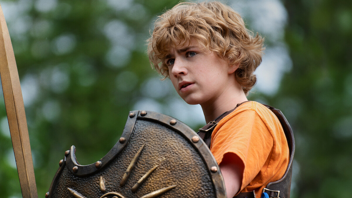 A young person with curly blonde hair in an orange shirt and armor holds a decorated shield outdoors, surrounded by green trees and natural light, appearing alert and determined.