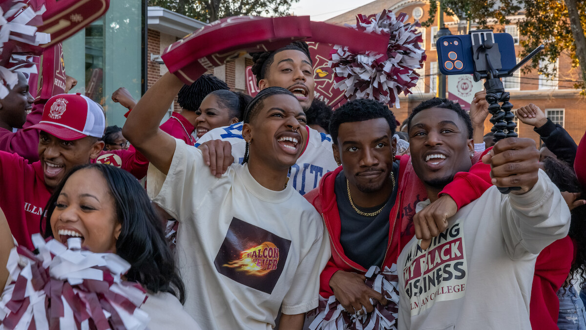 Grupo de estudantes sorrindo, comemorando e tirando uma selfie ao ar livre durante um evento animado no campus. Todos usam as cores da universidade e seguram pompons, com prédios históricos do campus ao fundo.