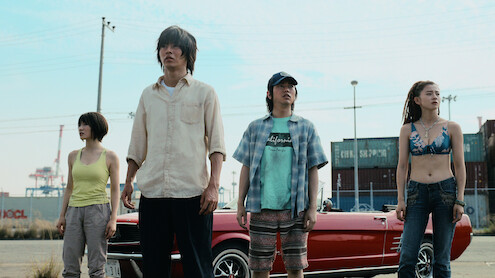 Four people stand in front of a red convertible car in an industrial area with shipping containers and power lines visible in the background, under a clear sky.