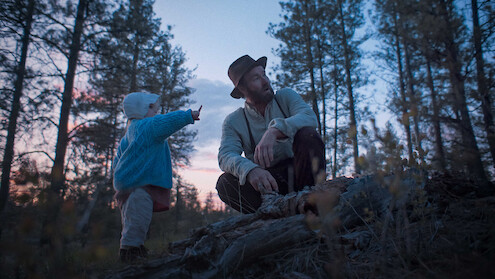 A man in a hat and a child in a blue sweater explore a forest at dusk, surrounded by tall trees. The child points at something, creating a sense of curiosity and peacefulness in the natural setting.