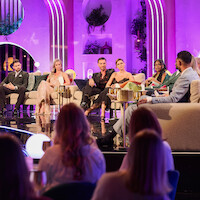 A group of people sit on stage in a brightly lit talk show set with purple lighting, decorative plants, and a live studio audience watching.